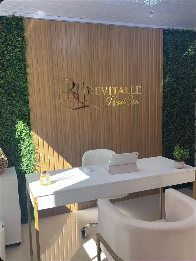 Recepção da Revitalle Head Spa: painel de madeira ripada com o logo dourado, paredes verdes, mesa branca com pés dourados e poltrona creme